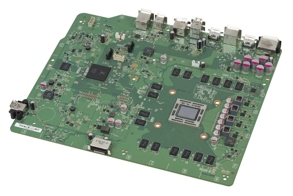 Scheda madre di una Xbox One vista di tre quarti: il processore al centro, le memorie intorno e le porte sul lato lungo
